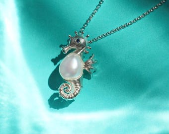 May include: A silver seahorse pendant necklace with a large white pearl belly and a small blue gem eye. The necklace is on a silver chain, and the background is a teal fabric. The text "AFRA JEWELRY STUDIO" is at the bottom.