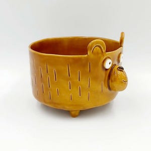 Può includere: Un vaso di ceramica a forma di orso, con finitura marrone dorato lucido. Il vaso ha un corpo rotondo, piedini e una faccia da orso con occhi bianchi e bocca sorridente. Linee verticali decorano il corpo.