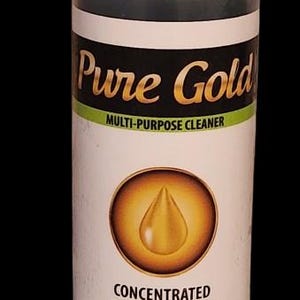 May include: A white plastic bottle with a black label that says "Pure Gold Multi-Purpose Cleaner". The label also has a gold circle with a yellow drop of liquid inside. The text "Concentrated Biodegradable" is printed below the circle. The bottle is sitting on a black surface.