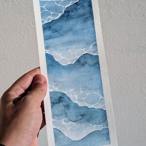 May include: Watercolor bookmark depicting an abstract ocean scene. The artwork uses shades of blue with white accents, creating a visual representation of waves. The bookmark is rectangular with a white border.