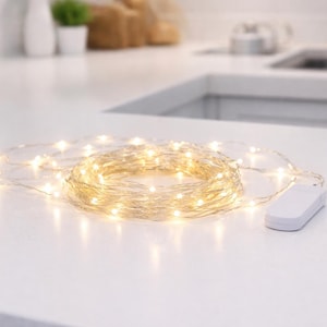 May include: A string of warm white LED fairy lights coiled on a white surface. The lights are connected to a small white battery pack. The lights are a decorative element for home decor and events.