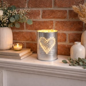 May include: A cylindrical silver metal tin lantern featuring a perforated heart design on the front. The interior is illuminated by warm string lights that shine through the small holes, creating a glowing heart shape on the metallic surface.