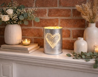 May include: A cylindrical silver metal tin lantern featuring a perforated heart design on the front. The interior is illuminated by warm string lights that shine through the small holes, creating a glowing heart shape on the metallic surface.