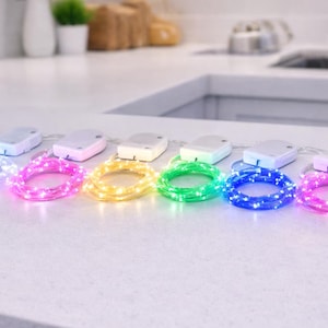 May include: A collection of battery-powered string lights in various colors, including white, pink, yellow, green, blue, and purple. Each light strand is coiled and connected to a small, white battery pack. The lights are arranged on a white countertop.