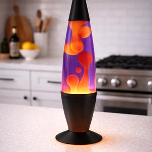 May include: A lava lamp with a black base and top, and a purple and orange liquid interior. The lamp is on a white countertop in a kitchen setting. The lamp's base is approximately 6 inches in diameter.