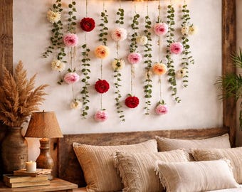 May include: A wall hanging featuring a horizontal wooden branch wrapped in warm white string lights. Multiple vertical strands of green eucalyptus leaves are suspended from the branch, interspersed with artificial pink, red, and cream flowers.