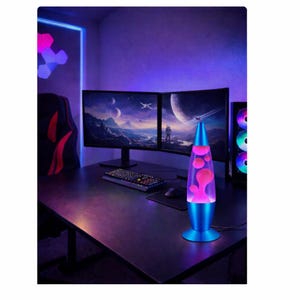 May include: A blue lava lamp with pink wax is on a black desk. The lamp is in front of two computer monitors displaying a space scene. The room has purple lighting and a gaming setup.