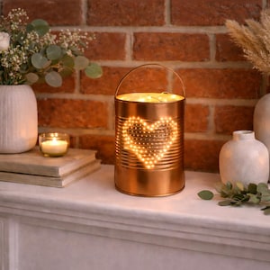 May include: A copper-colored lantern with a heart-shaped light display, surrounded by candles, vases, and flowers. The lantern is on a white mantel against a brick wall, creating a warm, inviting ambiance.