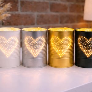 May include: Four decorative tin can lanterns in white, silver, gold, and black. Each lantern features a heart-shaped design made of small, warm white lights. The cans are arranged on a white surface.