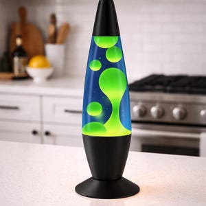 May include: A black lava lamp with a blue and green liquid inside. The lamp has a black base and top, and the liquid is a bright green color. The lamp is on a white countertop in a kitchen setting.