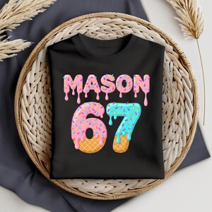 Può includere: T-shirt nero con il nome "MASON" in lettere rosa gocciolanti e il numero "67" in un design a cono gelato rosa e blu. La maglietta è presentata in un cestino di vimini.