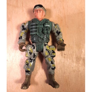 Puede incluir: Figura de soldado de juguete de plástico en posición de pie. La figura viste un uniforme de camuflaje con detalles verdes y amarillos. Tiene casco, chaleco y botas. El juguete está sobre una superficie de madera marrón claro.