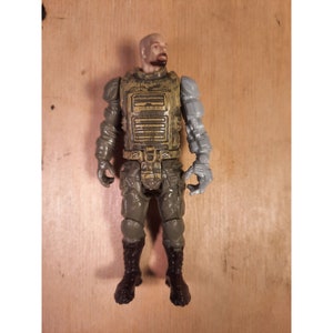 Peut inclure: Une figurine d'action verte et brune avec un bras robotique gris. La figurine a une barbe et porte une armure détaillée.