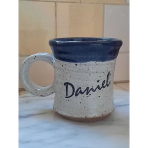 May include: A handmade ceramic mug with a speckled white body and a dark blue rim. The name "Daniel" is written in black script on the front. The mug has a large, rounded handle.