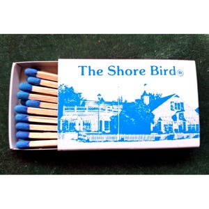 May include: A white matchbox with blue text and a blue illustration of a building with the text "The Shore Bird®" on the front.