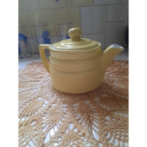 May include: A yellow ceramic teapot with a lid and a handle. The teapot is sitting on a crocheted doily.