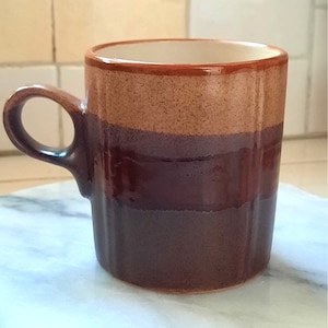 May include: A brown and tan ceramic mug with a wide handle. The mug has a brown band around the middle and a tan band around the top.