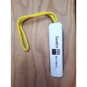 May include: White plastic key chain with a yellow lanyard. The keychain has the text "Sandy's Ski & Sports" printed on it.