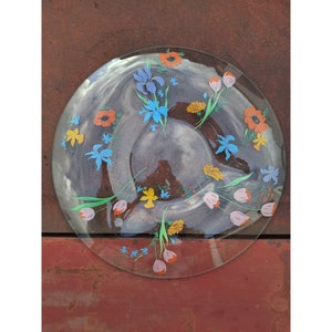 May include: A clear glass serving dish with a floral design. The flowers are blue, yellow, orange, and pink. The dish is round and has a slightly raised rim.