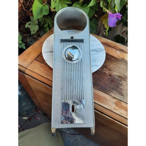 May include: A vintage silver-toned metal object with a circular opening at the top and a rectangular body. The body features vertical grooves and a circular, reflective element. The object rests on a white marble surface.