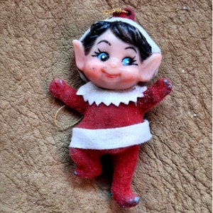 May include: A vintage Christmas ornament featuring a smiling elf with black hair, blue eyes, and a red and white outfit. The elf is wearing a red felt suit with a white trim and a white felt hat.