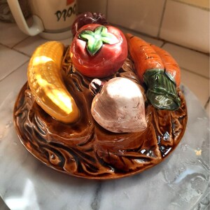 May include: A ceramic dish with a brown glaze features a stylized arrangement of vegetables. The dish includes a yellow corn cob, a red tomato, a white onion, and a green carrot.