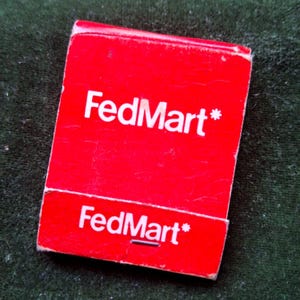 Pode incluir: Um maço de fósforos vermelho com o texto "FedMart*" em branco, repetido na parte superior e inferior. O maço de fósforos é retangular e tem uma aparência desgastada.