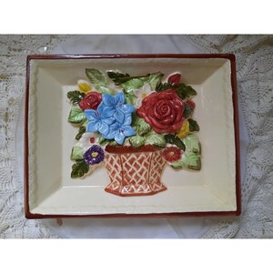 May include: A ceramic rectangular serving tray with a raised floral design. The tray features a basket of flowers with red, blue, yellow, and white flowers. The tray has a brown border.