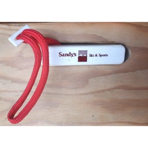 May include: A white plastic key chain with a red lanyard. The key chain has the text "Sandy's Ski & Sports" in red lettering.