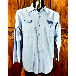 May include: Light blue long-sleeve button-up shirt with two front pockets. The shirt has a patch that says "Disneyland" and another patch that says "Gus". The shirt has black buttons and some white paint marks.