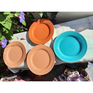 Puede incluir: Cuatro platos de cerámica en diferentes colores: naranja, marrón claro, marrón claro y azul. Los platos están dispuestos sobre una superficie blanca.