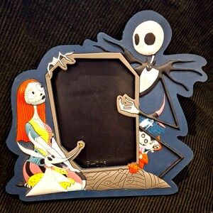 May include: A blue-edged picture frame featuring characters from The Nightmare Before Christmas. Jack Skellington, Sally, and Zero are depicted, with a black frame center. The frame has a gray tombstone design at the bottom and a dark background.