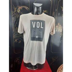 May include: A beige t-shirt with a black and white graphic print of the word "VOLCOM" in a box. The graphic also includes the words "PRESERVE DISCOVER" and a small logo.