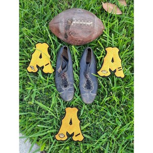 May include: A brown leather football with white stitching and the text "Rawlings" on it.  Four yellow felt letters spelling "AMA" are arranged around the football. Two black leather shoes with laces are on either side of the football.