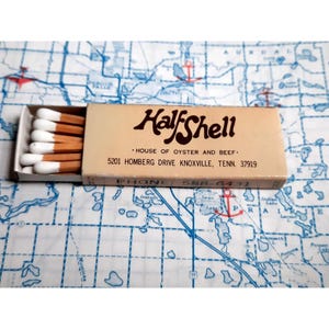 May include: A matchbook with a white background and brown text. The text reads "Half Shell - House of Oyster and Beef. 5201 Homberg Drive Knoxville, Tenn. 37919. Phone 588-6431."