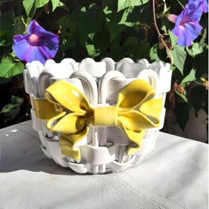 May include: White ceramic basket with a yellow bow. The basket is woven and has a decorative design.