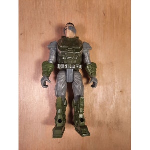Peut inclure: Une figurine d'action grise et verte avec un casque et un fusil. La figurine est debout sur une surface en bois.