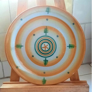May include: A round ceramic plate with a concentric circle design in shades of orange, white, and green. The center of the plate features a stylized star with four points.