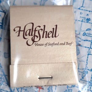 May include: A vintage matchbook cover with a white background and brown text. The text reads "Halfshell House of Seafood and Beef".