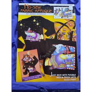 Könnte beinhalten: Ein schwarzes Langarmshirt mit einer nähfreien Stoffapplikation einer Hexe, die auf einem Besen fliegt. Die Applikation ist von Sternen und einem Halbmond umgeben. Das Shirt ist Teil eines Bastelsets, das Anweisungen zum Anbringen der Applikation mit Fusible Web und Slick Paint enthält. Das Set heißt "No-Sew Fabric Applique" von Daisy Kingdom.