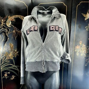 May include: A gray zip-up sweatshirt with the text "UGC COM" in a pink and black design. The sweatshirt has a collared neckline and ribbed cuffs and hem. The zipper runs down the center of the garment.