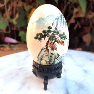 May include: A hand-painted egg with a landscape scene featuring a pine tree, mountains, and a small village. The egg is displayed on a black stand with three legs.