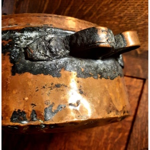 May include: A vintage copper pot with a black handle. The pot has a worn, aged look and shows signs of use.