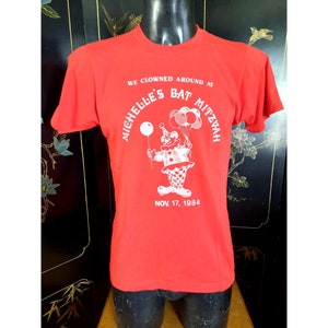 May include: Red t-shirt with a white graphic of a clown holding balloons. The text on the shirt reads "We Clowns Around At Michelle's Bat Mitzvah Nov. 17, 1984".
