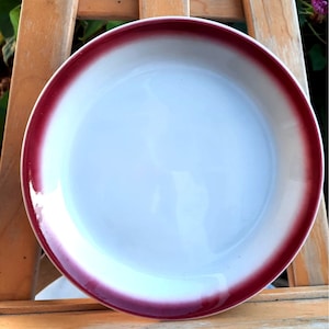 May include: A white ceramic plate with a wide, dark red rim. The plate has a slight gradient in the red rim, fading to a lighter shade of red.