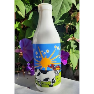 Puede incluir: Una botella de leche de vidrio blanco con una etiqueta azul, verde y amarilla que presenta una vaca de dibujos animados con un sombrero rojo y un sol con un centro amarillo y rayos blancos. La etiqueta también tiene margaritas blancas.