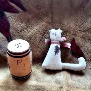 May include: A white fabric cat with a red and white plaid bow and a brown tail. The cat is sitting on a brown surface. There is a wooden salt and pepper shaker with the letter "P" on it in the foreground.