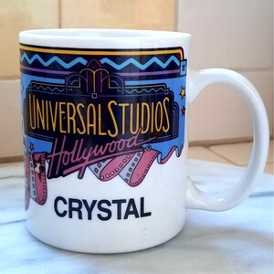 Puede incluir: Taza de cerámica blanca con un diseño azul, amarillo y rosa con el texto "Universal Studios Hollywood" y "Crystal".