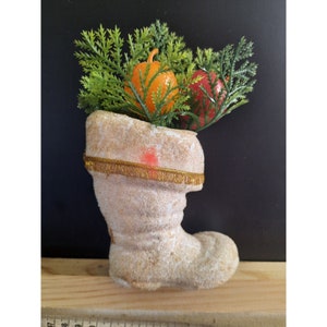 May include: A white, snow-covered boot with a gold trim, filled with a green plant and two orange and red candles.