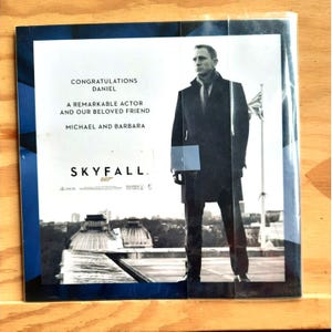 Op de afbeelding: Een zwart-wit poster voor de film Skyfall, met Daniel Craig als James Bond. De poster heeft de tekst "Gefeliciteerd Daniel, Een opmerkelijke acteur en onze geliefde vriend, Michael en Barbara, Skyfall 007".